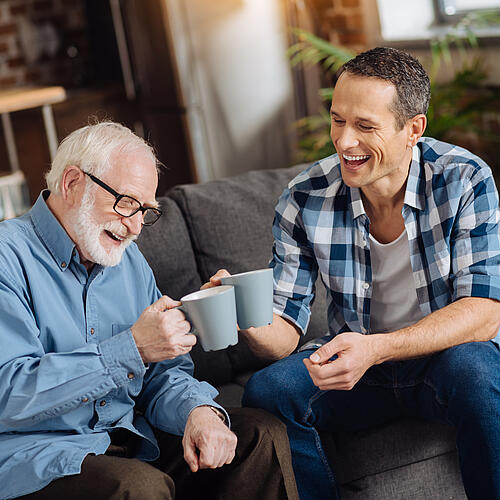 Ein jüngerer und ein älterer Mann sitzen auf einem Sofa und stoßen lachend mit ihren Bechern an.